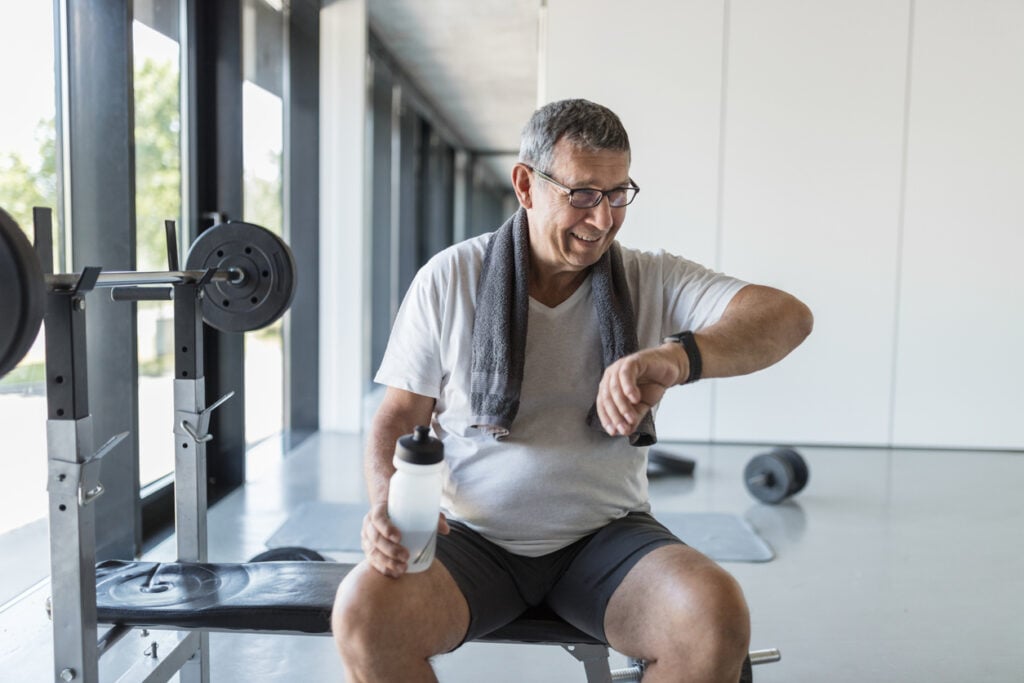 Gym interior, older man on bench center frame, midday natural light, documentary style, fitness props visible, one person.