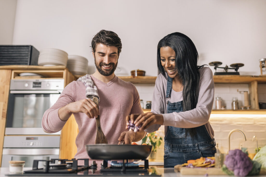 Modern kitchen, couple cooking together, woman adding veggies to pan, bright lighting, editorial travel photo, two people.