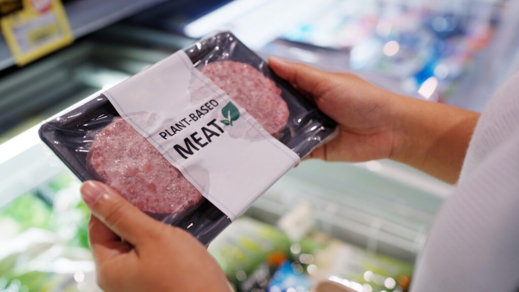 Grocery store refrigerated aisle, package of Plant-Based Meat in hand, centered view, bright indoor lights, documentary style, person present.