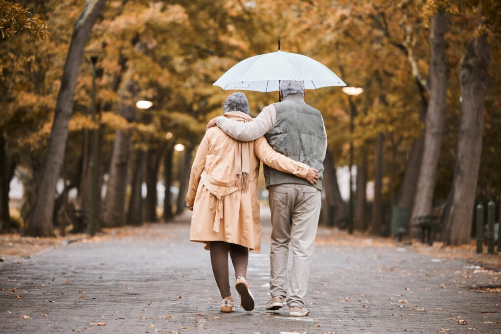 Tree-lined park path, elderly couple under blue umbrella, autumn leaves scattered, overcast sky, editorial travel photo, people.