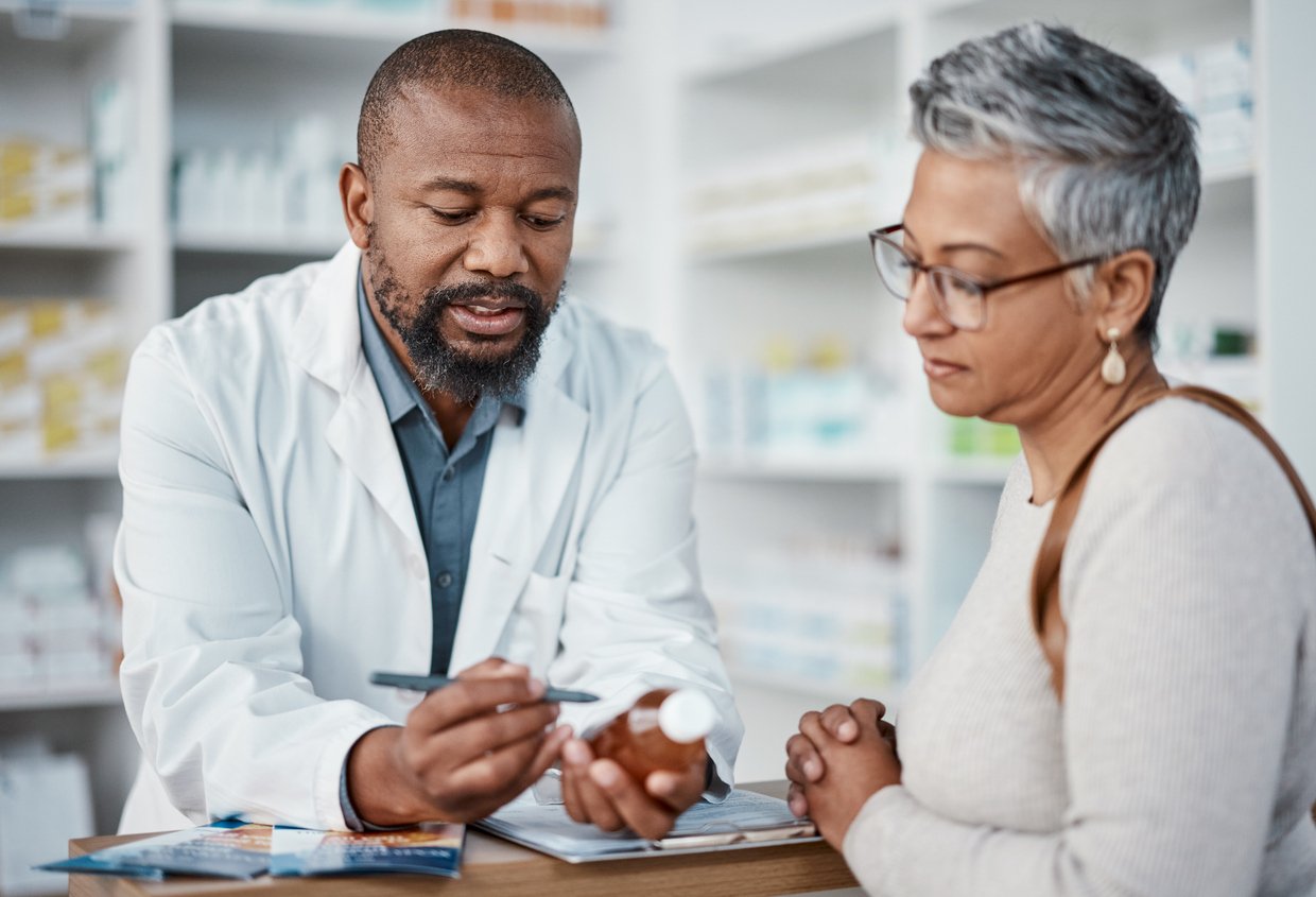Pharmacy, pharmacist explains meds to woman, mid-shot, daytime fluorescent light, editorial travel photo, two people.