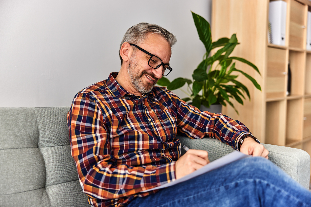 Cozy living room, man in plaid shirt writing, centered mid-shot, daytime natural light, documentary style, one person.