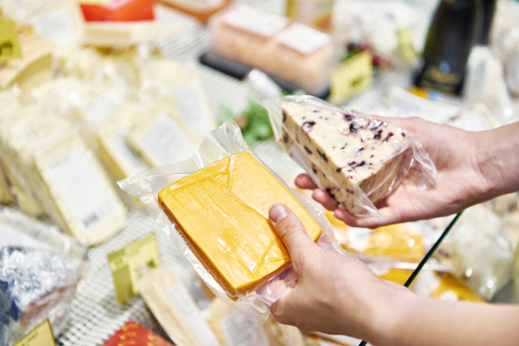 Cheese store, packaged cheeses in hands, overhead shot of variety display, daytime ambient light, documentary style, one person.
