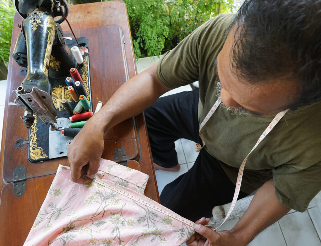 Wooden table workspace, tailor measuring floral fabric, sewing machine and spools foreground, soft daylight, documentary style, one person.