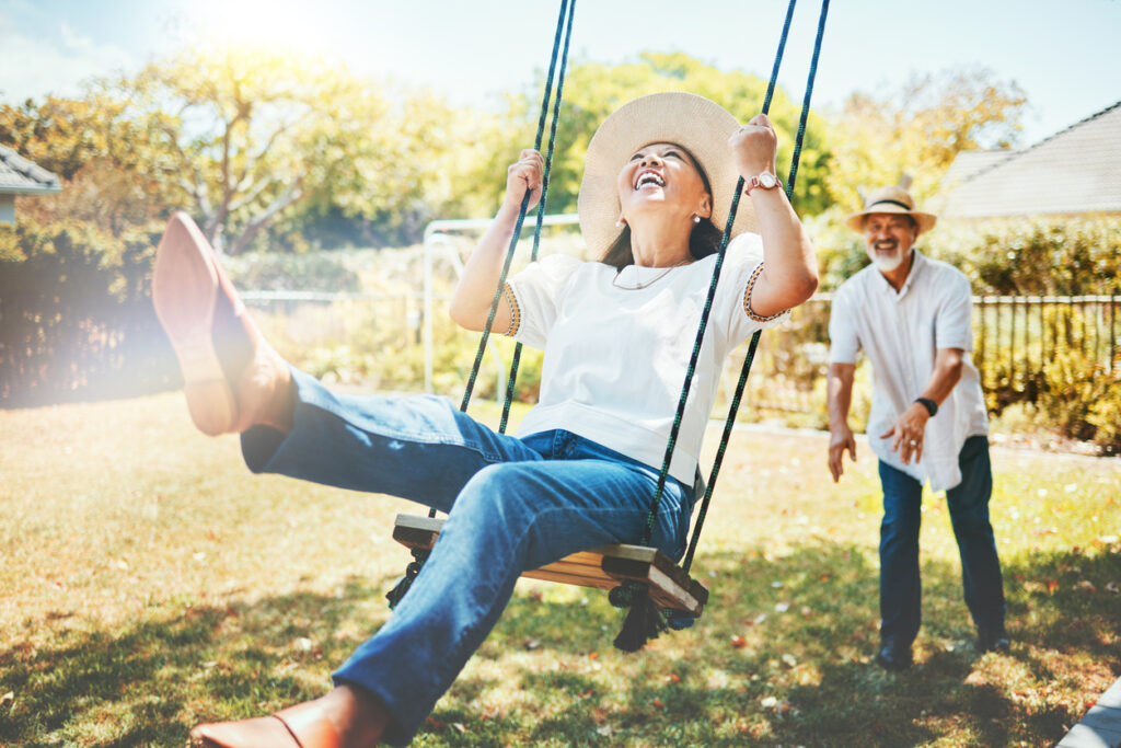 Sunny backyard, woman mid-swing foreground, man behind pushing, morning golden light, editorial travel photo, two older adults.