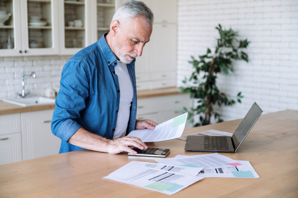 Kitchen interior, older man with calculator, documents and laptop scattered, daytime natural light, documentary style, one person.