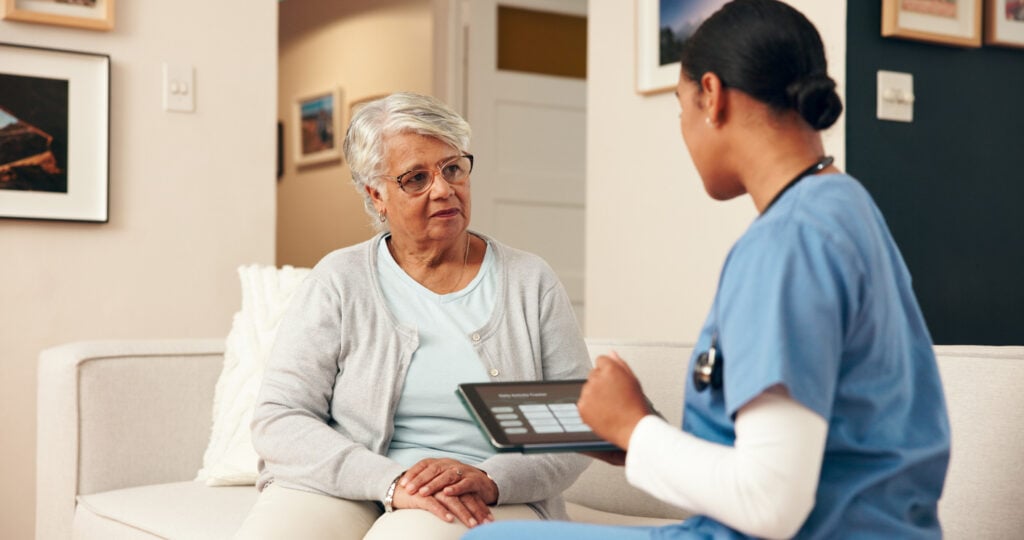 Home interior, nurse with tablet, seated on couch beside elderly woman, soft natural light, documentary style, two people.
