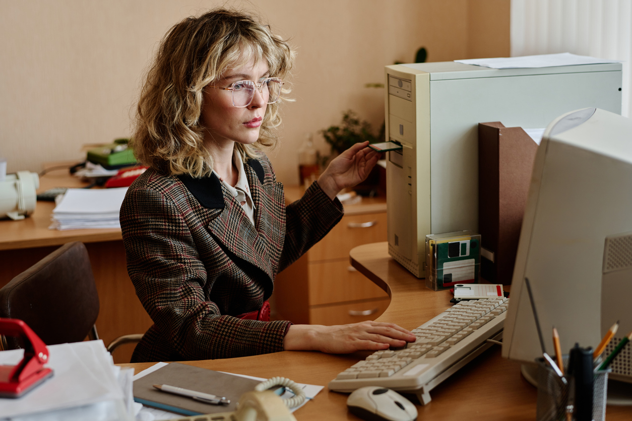 Office desk, woman with glasses, vintage computer and floppy disk, papers scattered, soft daylight, documentary style, people.