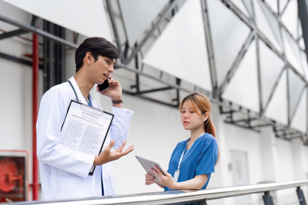 Modern building exterior, male doctor on phone with clipboard, side by side nurse with tablet, daytime, editorial travel photo, people.