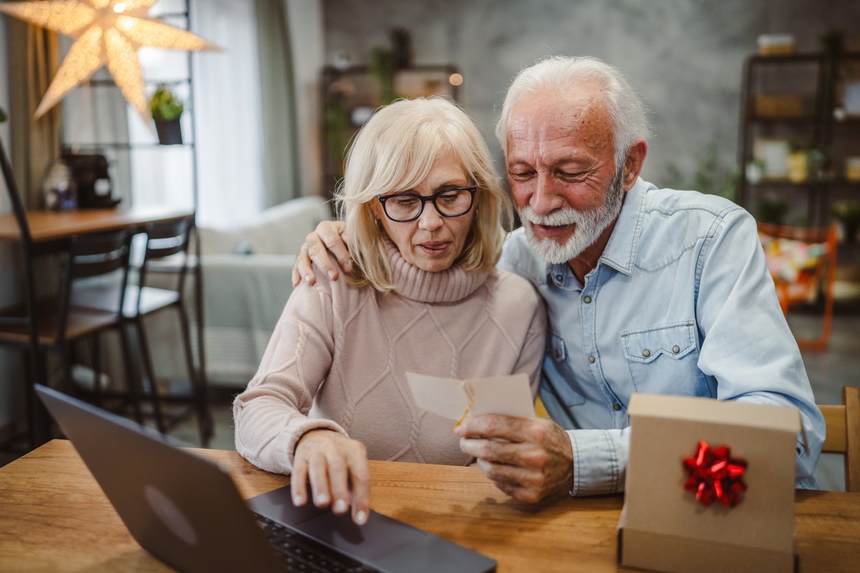 Kitchen table, elderly couple sharing photo, close-up with laptop and gift box, daylight, documentary style, two people.