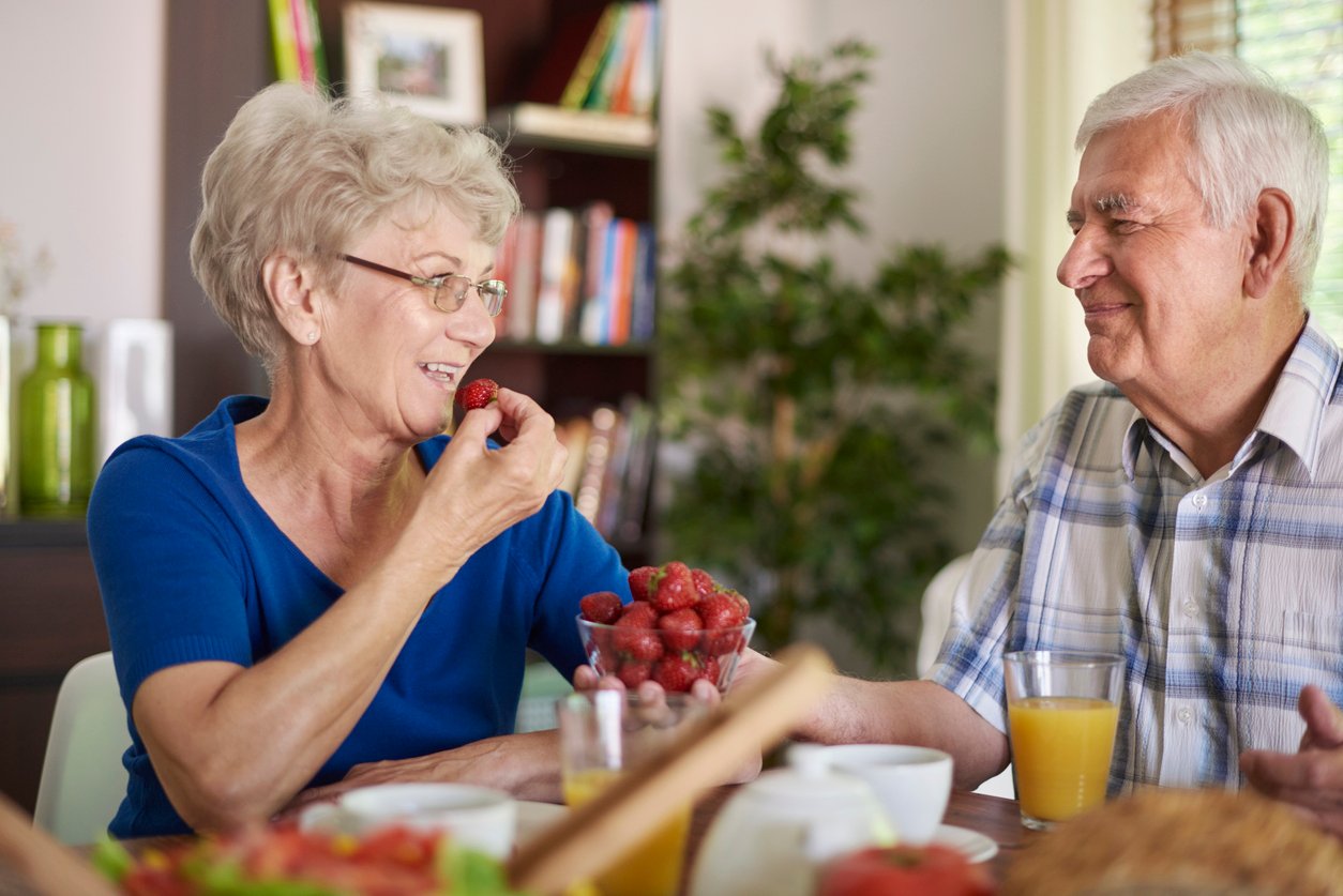 Dining room, smiling elderly woman eating strawberry, shared table with meal, bright daylight, documentary style, two people.