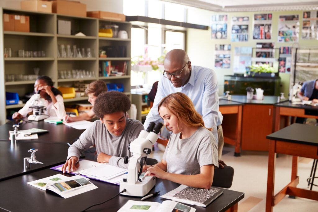Science classroom, students using microscopes, group at lab tables, bright indoor lighting, documentary style, teacher and students.
