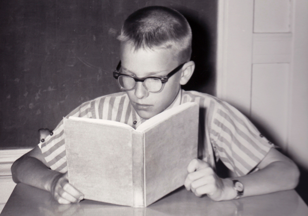 Classroom, boy reading at desk, glasses and striped shirt, natural daylight, documentary style, one child present.