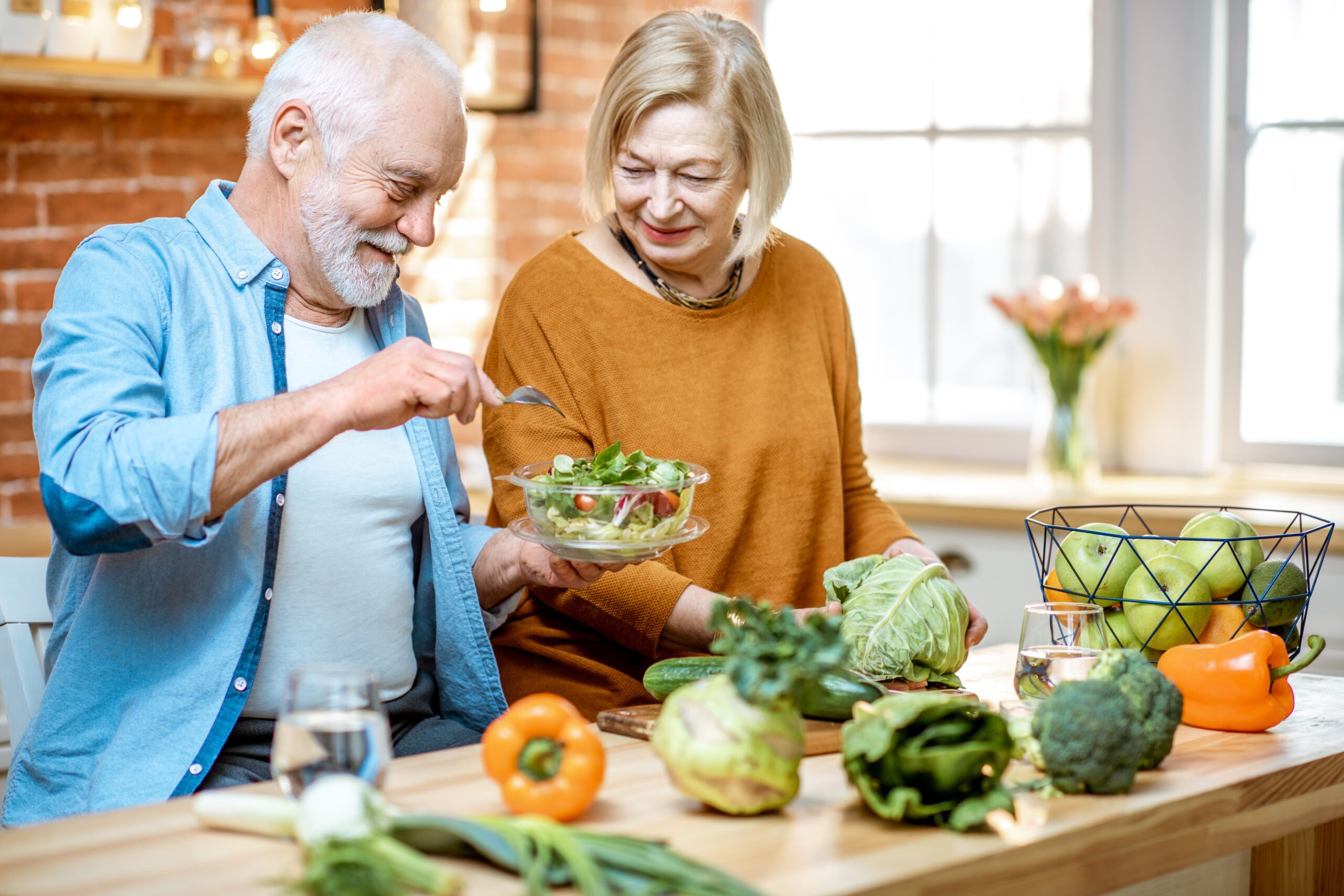 Bright kitchen, elderly couple making salad, colorful vegetable spread foreground, daylight, documentary style, two people.