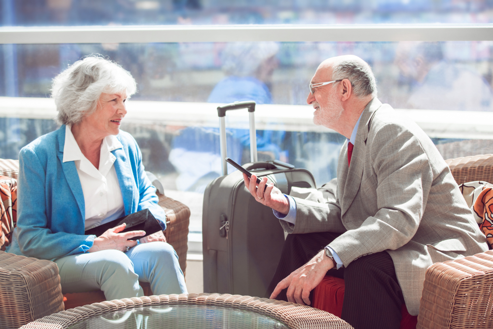 Airport terminal, wicker chairs and suitcase foreground, older couple talking, sunlight streams through windows, editorial travel photo, with people.