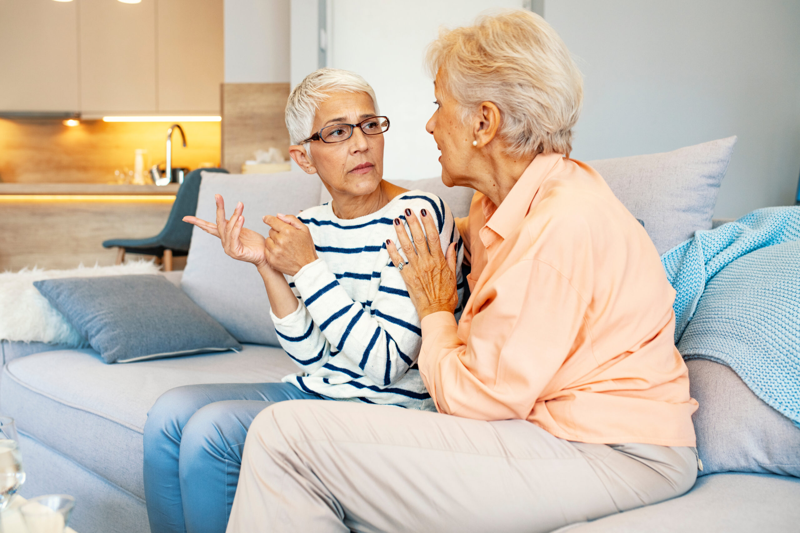 Living room, two women talking, couch center-frame, soft daylight, documentary style, people.