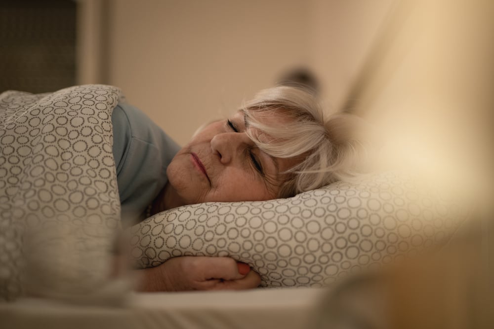 Bedroom, older woman sleeping, patterned bedding framing scene, soft ambient lighting, editorial travel photo, people present.