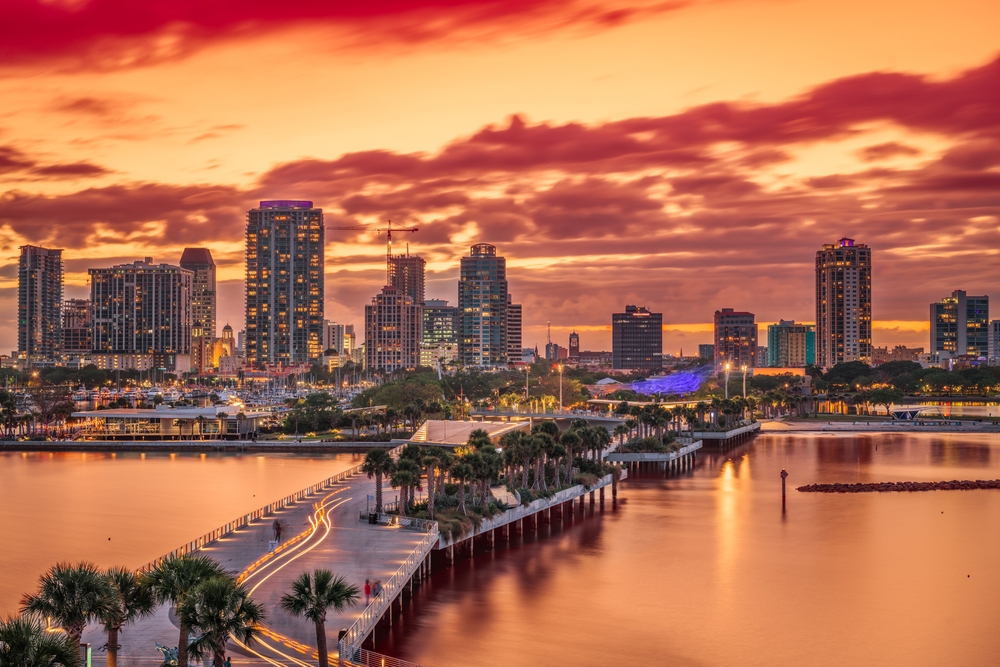 Downtown city skyline, tall buildings, orange sunset reflection, waterfront road with palm trees, editorial travel photo, no people.