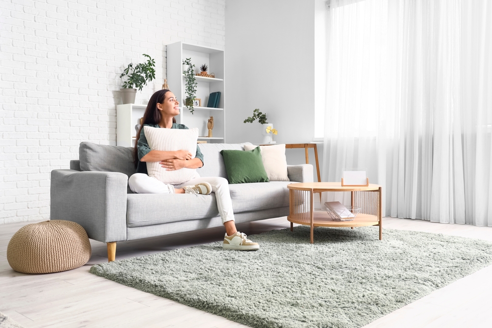 Modern living room, woman hugs pillow on sofa, window view, bright daylight, editorial home photo, single person.