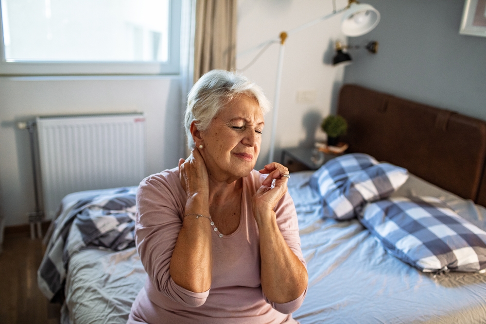 Bedroom interior, elderly woman at bedside rubbing neck, soft ambient light, documentary style, one person present.