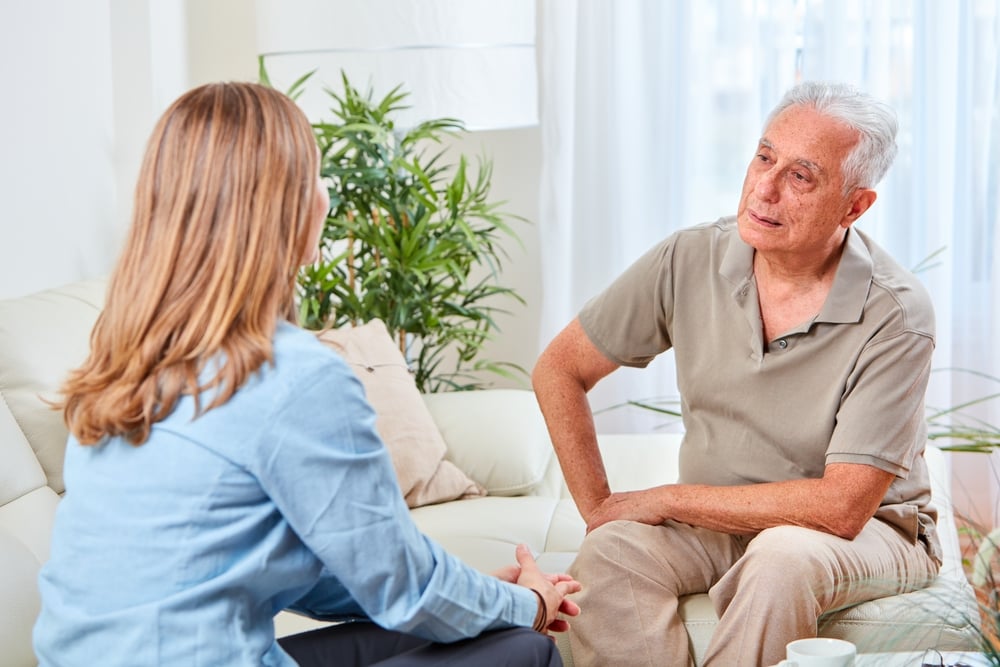 Living room, older man and woman talking, side view on couch, daylight through curtains, documentary style, two people.