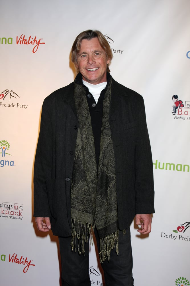 Event backdrop with logos, blond man in black coat and green scarf centered, indoor lighting, editorial photo, people.
