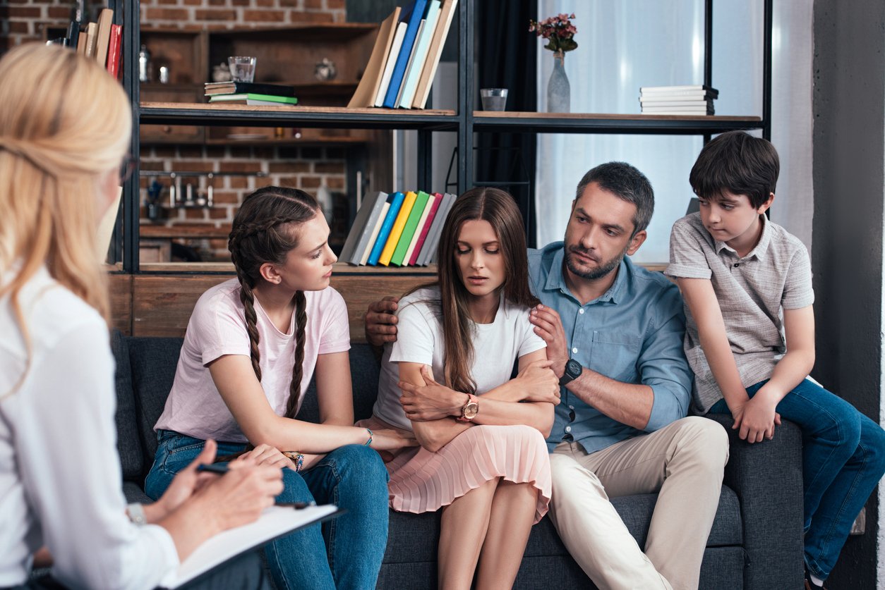 Modern therapy room, distressed woman comforted by family, group seated in circle, natural light, documentary style, people.
