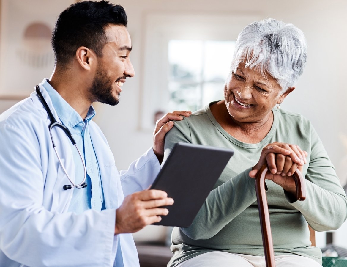 Bright medical office, doctor with tablet, seated next to elderly woman with cane, daytime light, editorial travel photo, people.