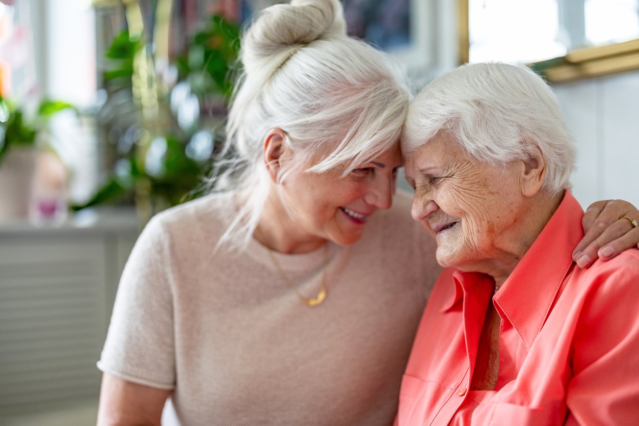 Cozy indoor room, two smiling elderly women, close-up foreground, soft evening light, documentary style, people present.