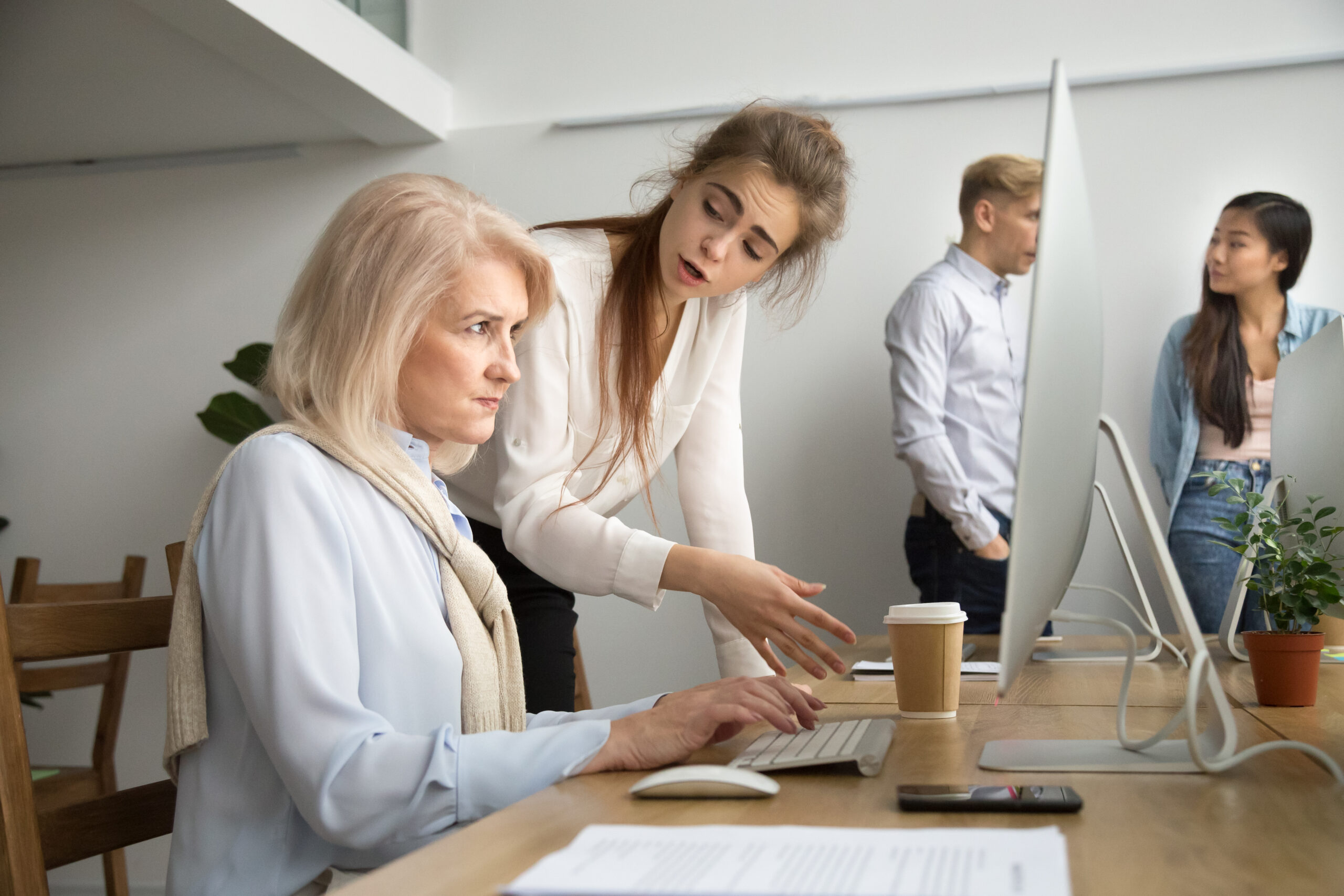Modern office, women collaborate at shared desk, foreground focus, afternoon light, documentary style, four people present.