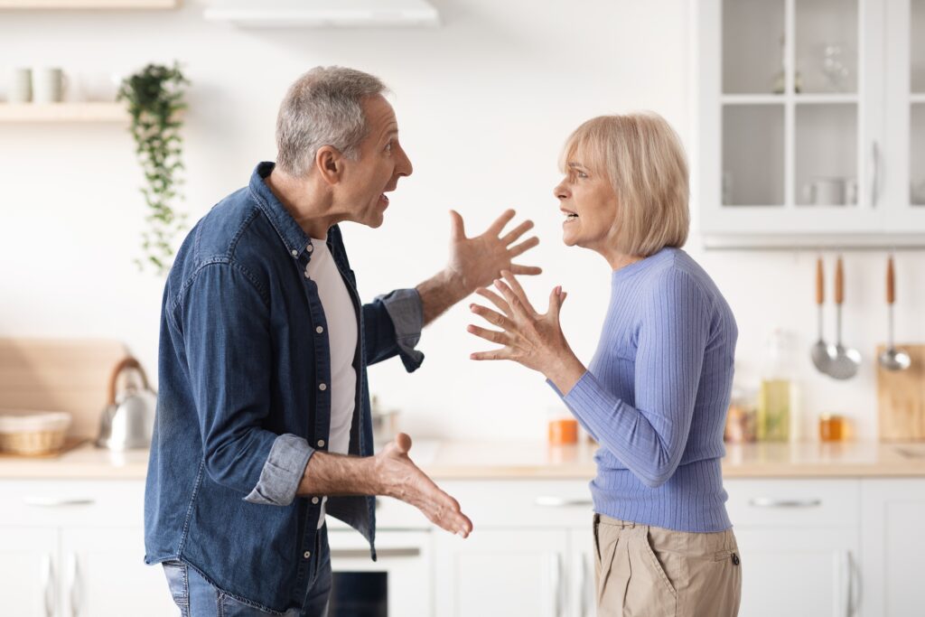 Kitchen, elderly couple arguing, facing each other with raised hands, bright modern space, documentary style, two people.