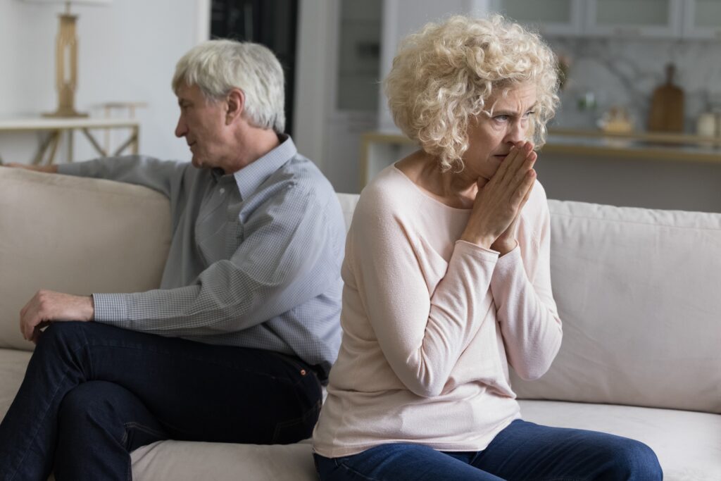 Living room, older couple back-to-back on couch, tense mood, soft indoor light, documentary style, two people.