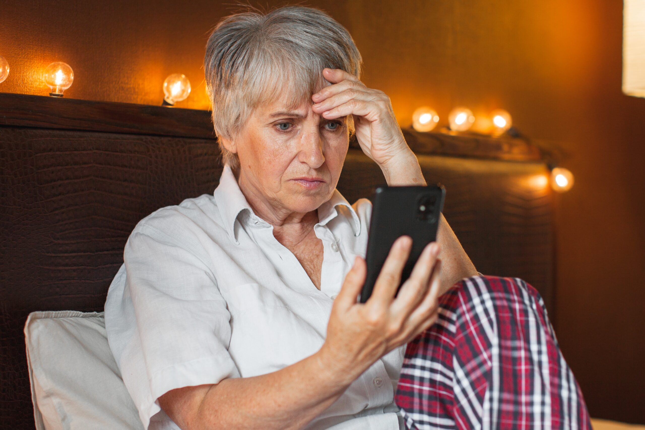 Bedroom, gray-haired woman on bed foreground, holding phone, evening with string lights glowing, documentary style, people.