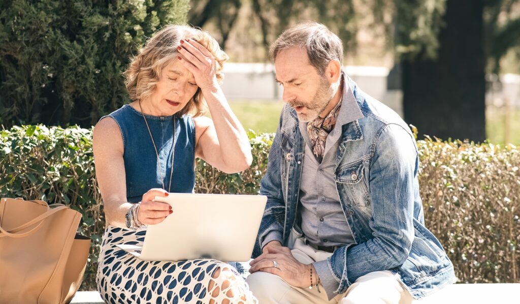 Park bench, elderly couple worried over laptop, side view, midday natural light, documentary style, people.